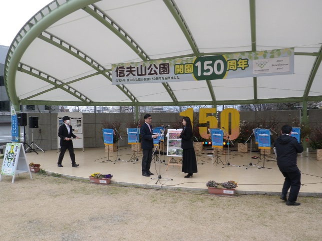 「信夫公園開園150周年」と書かれた横断幕と「150」と書かれたパネルが設置されているステージ上に関係者2名が向き合っている様子の写真