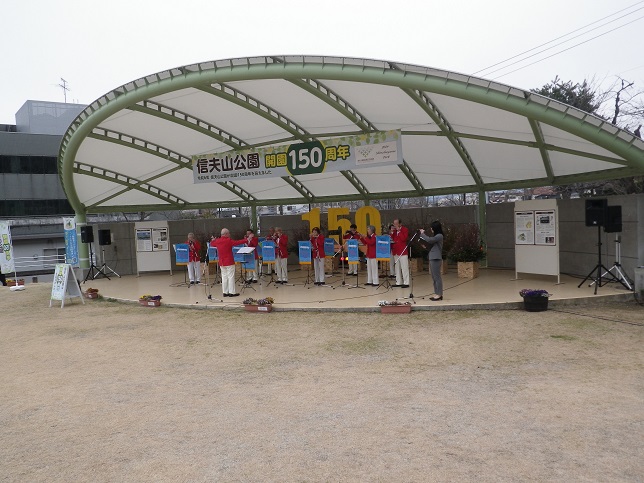 「信夫山公園開園150周年」と書かれた横断幕が設置されたステー上で赤色のジャケットを着た関係者が演奏を行っている様子の写真
