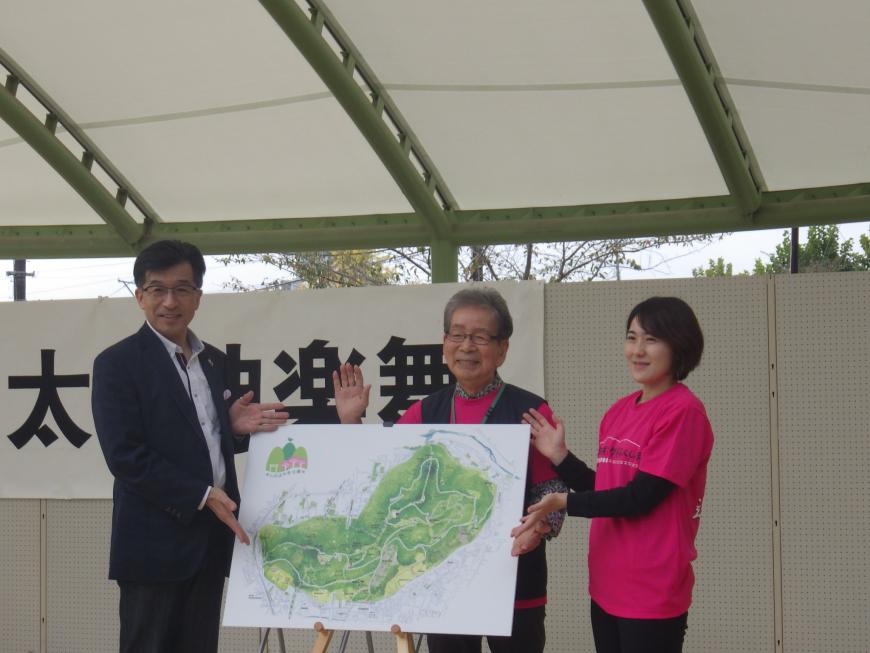 ステージ上でイーゼルに置かれた信夫山のマップを中心に市長と同じピンク色のTシャツを着た関係者がマップを披露するように両手を広げている写真