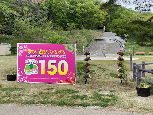 芝生の広場に信夫山公園開園150周年の記念のパネルと、その隣に赤や白などの花で装飾された花のガーデンアーチが設置されている写真撮影スポットの写真