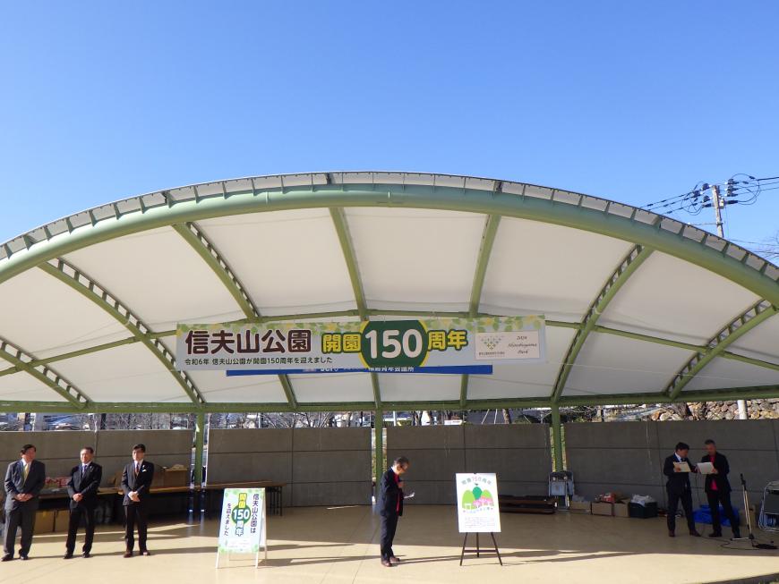 信夫山公園開園150周年と書かれた横断幕が設置されたステージ上でスーツ姿の関係者数名が立っている写真