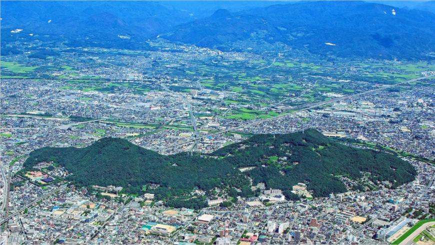 周りを住宅や建物が立ち並ぶ福島盆地の中央に緑色の染まった山々が連なってできた信夫山を上空から撮影した写真