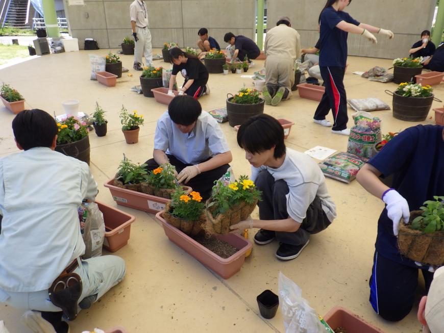 プランターに花を植えている作業服を着た関係者と2名の学生の写真