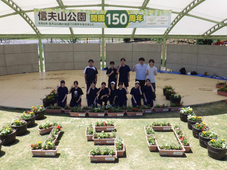 「信夫山公園開園150周年」と書かれた横断幕を背景に「150」という文字を造った花が植えられたプランターの前で学生たちが2列に並び記念撮影を行っている写真