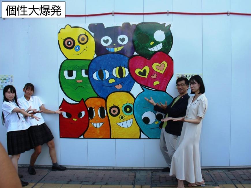 10個のキャラクターが描かれたアートの両横に両手のひらを上にあげ同じポーズをしている4名の写真