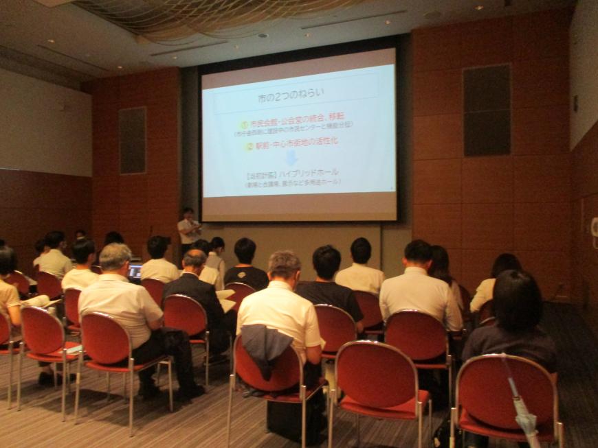 前方に資料が投影された大きなスクリーンがあり、参加者が椅子に座って話を聞いている会場を後方から撮影した写真