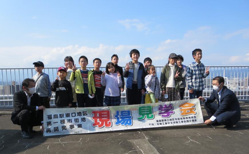 現場見学会の幕の後ろに参加した子供たちがピースをしながら並んで記念撮影をしている写真