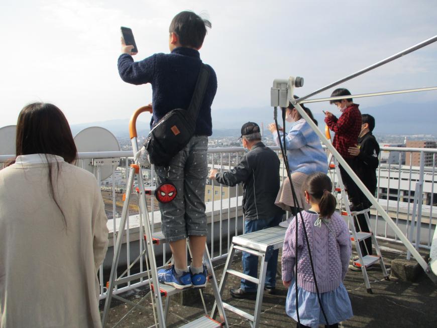 スマホを片手に3名の子供たちが脚立に乗りながら工事現場を写している写真