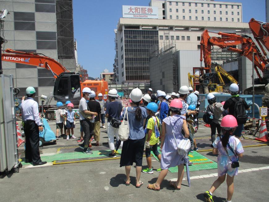 ショベルカーが置かれた工事現場をヘルメットを付けた参加者の人たちが見学をしている写真