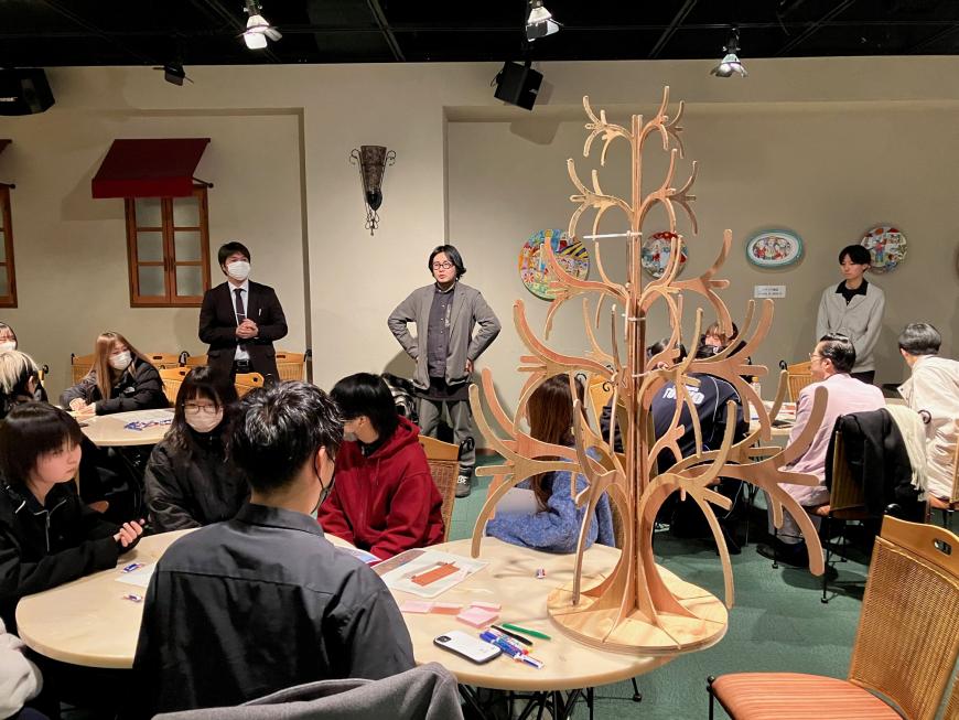 前方に立っている男性の話を、木で作られたツリーが飾られているテーブルの席に座った参加者の人たちが聞いている様子の写真