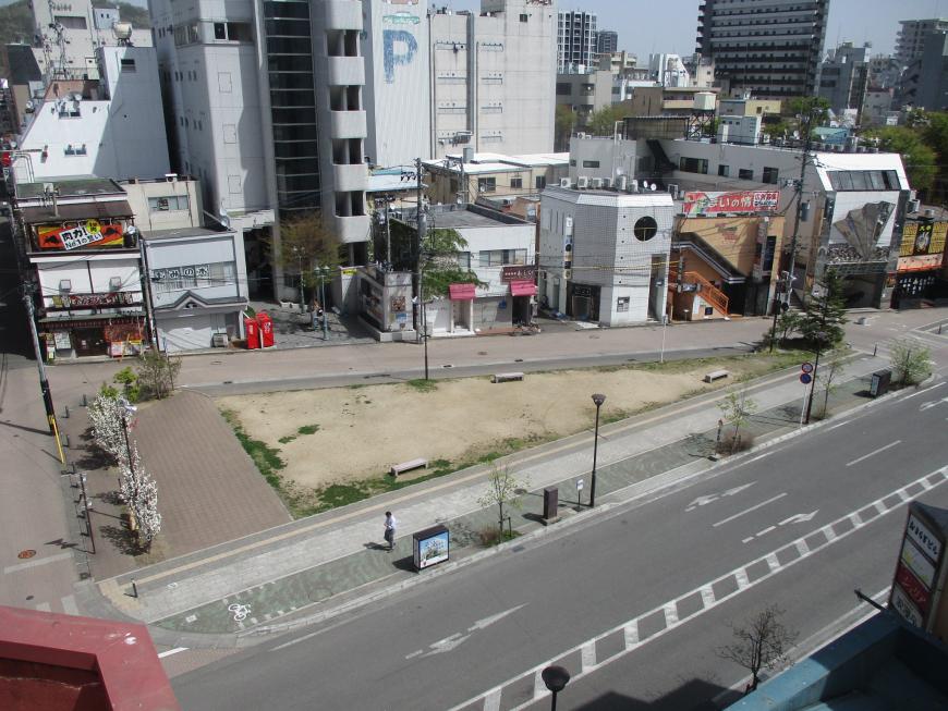 奥にビルや店舗などの建物があり、市道に囲まれて三角形の形をした広場の写真