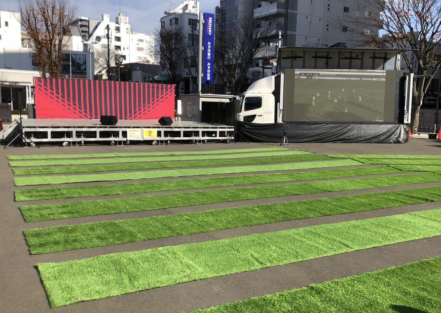 組み立て式のステージの横に大型ビジョンが搭載されたトラックが停まっており、観客席には人工芝マットが敷かれている写真