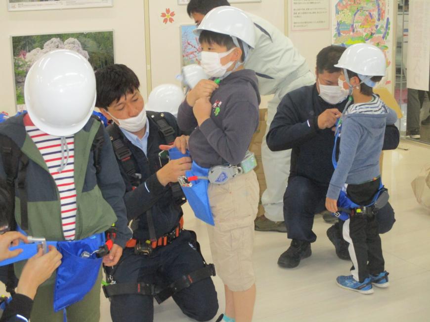 白いヘルメット姿の男の子3人が関係者の男性に青いベルトを着けてもらっている写真