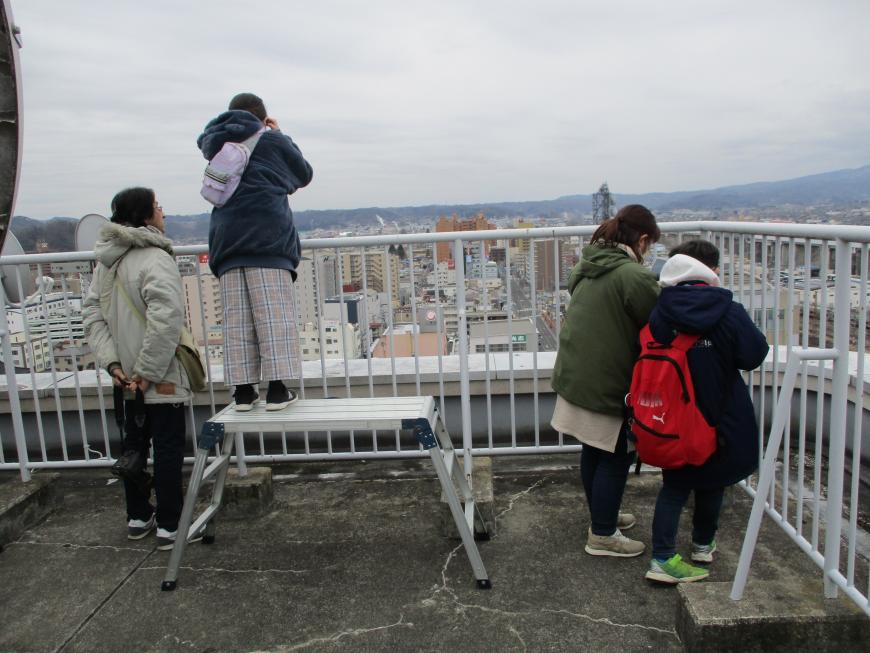 屋上jのフェンスの手前に設置された台の上に乗ったり、フェンスの間から街並みを撮影している2組を後方から写した写真