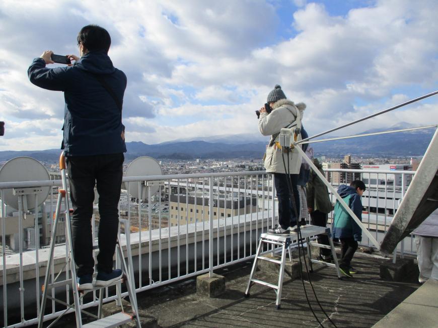 脚立に乗り2名の男女が工事現場を撮影している写真