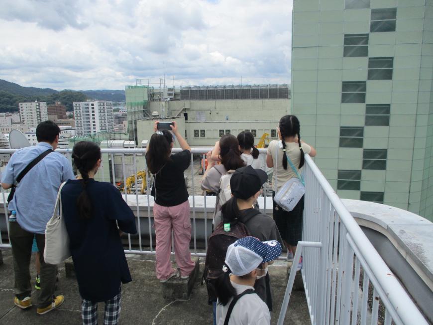 子どもたちがフェンスの土台にのりかかり工事の現場を撮影している様子を後方から写した写真