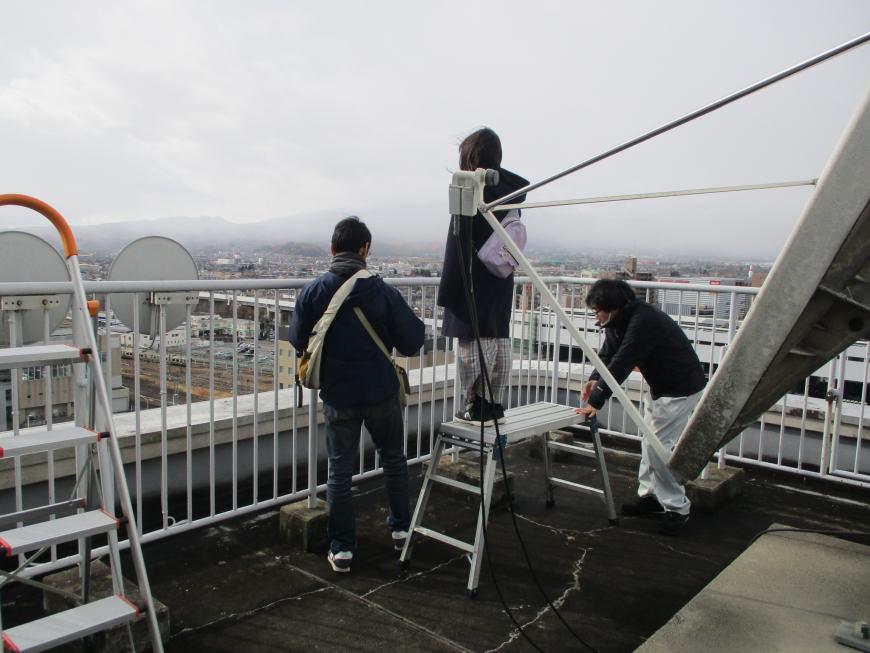 男性が抑えた脚立の上に女の子が真っすぐ立ち、男性がフェンス近くに立っている観測会の様子の写真