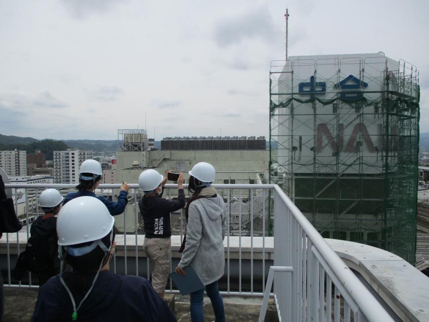 目の前で工事がされている建物をフェンス越しからヘルメット姿の子供たちが撮影をしている写真