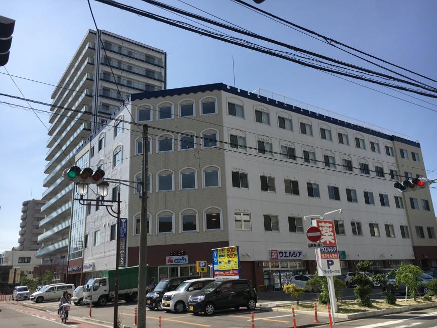 1階に薬局がある、白い外壁の5階建ての建物で目の前のコインパーキングに車が数台停まっている写真