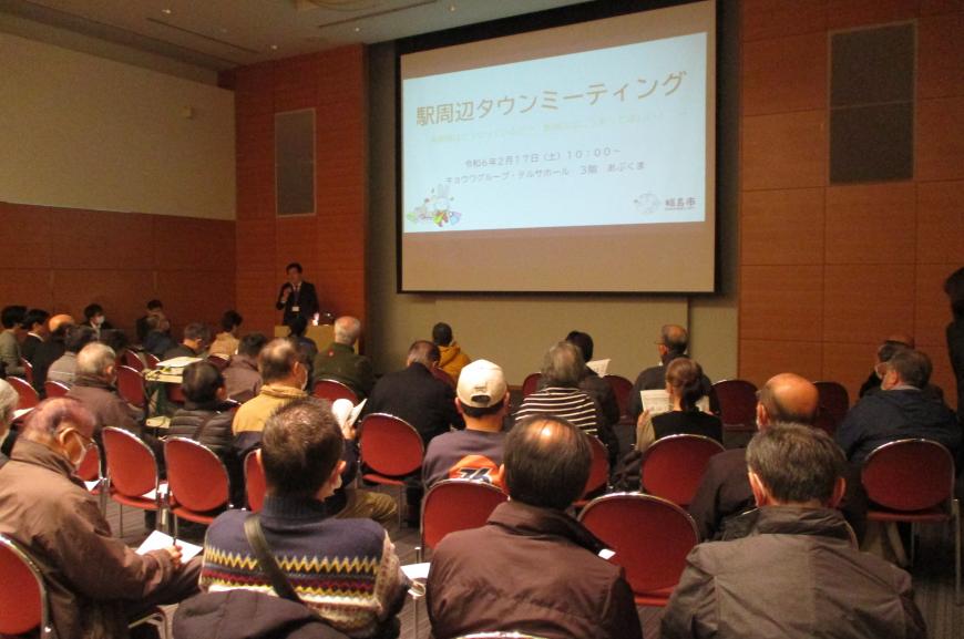 前方の資料が投影された大きなスクリーンの横で講師が説明をしており、大勢の参加者が椅子に座って話を聞いている会場を後方から撮影した写真
