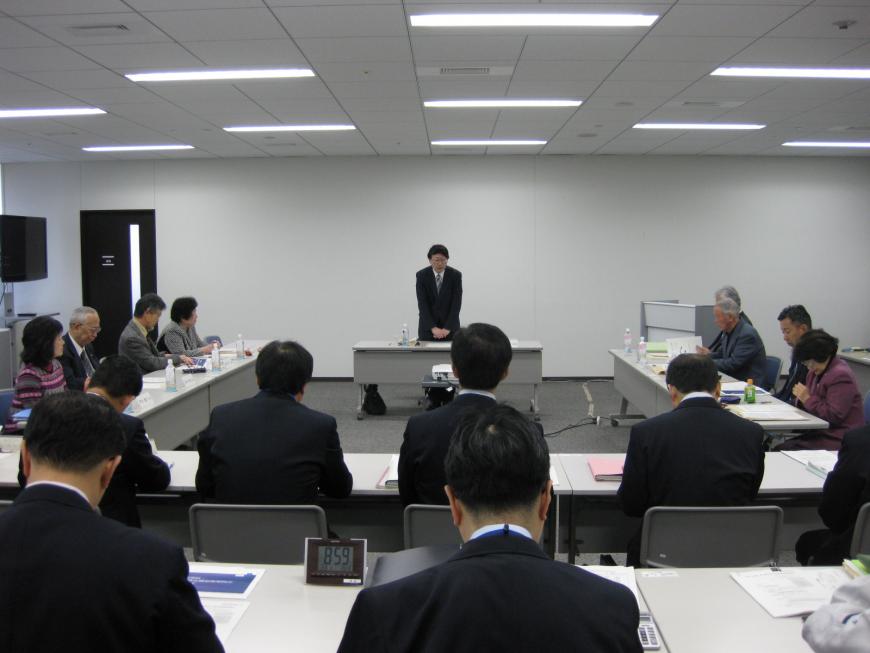 会場内に設置された長机の席に参加者の方々が座り、前方中央にて男性が席を立ち話をしている運営審議会の様子の写真