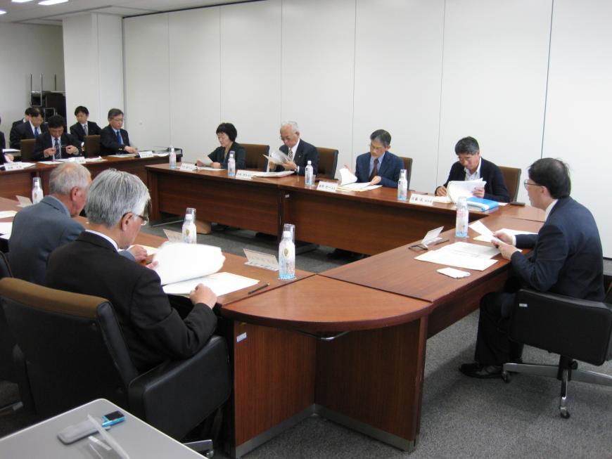 室内にロの字型に設置された机の席に関係者の方々が座り、審議会が行われている様子の写真