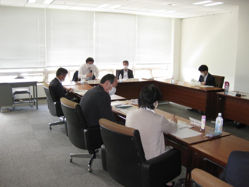 コの字型に設置されてた席に委員の方々が座っており、窓側に座っている男性がマイクを持って話をしている審議の様子の写真