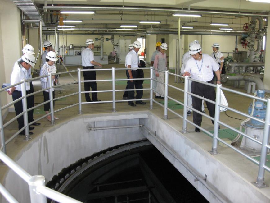 ヘルメットをかぶった関係者の方々が手すりのある高所から下に見える大きな処理施設を見下ろしている様子の写真