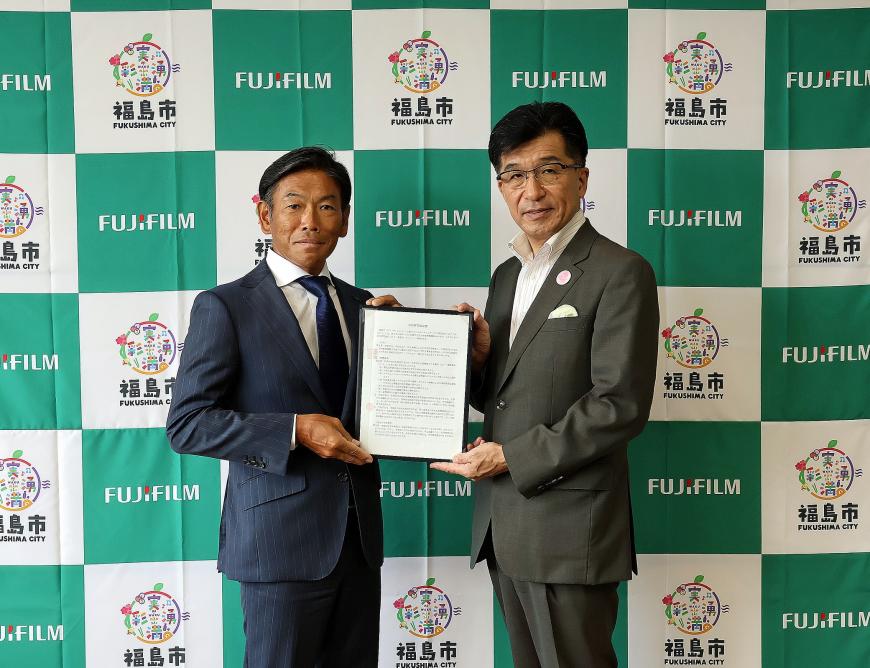 福島市のバックパネルの前で、代表の男性と市長が一緒に協定書を持ち並んでいる締結式の様子の写真