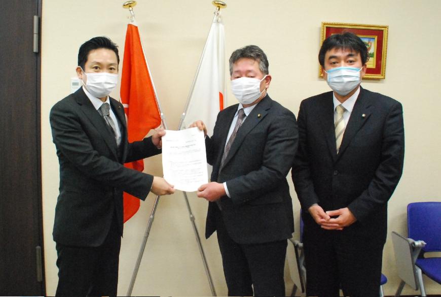 マスク姿の黒沢座長と真田議長が答申書をカメラの方を向け持ち、黒沢座長の横に立つ石原副座長と記念撮影している写真