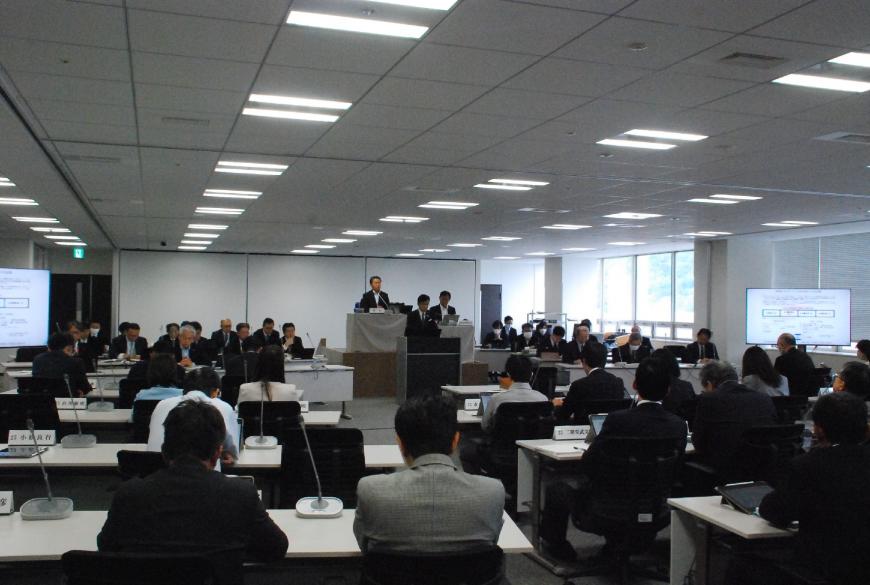 演台に男性が立ち、前方の左右のスクリーンには資料が映し出された協議会の様子の写真