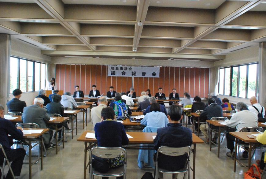 「議会報告会」の幕が吊るされた会場で長机が3列に並べられ、それに2人ずつ座っている30人ほどの参加者の前に7人の職員が座り報告をしている写真