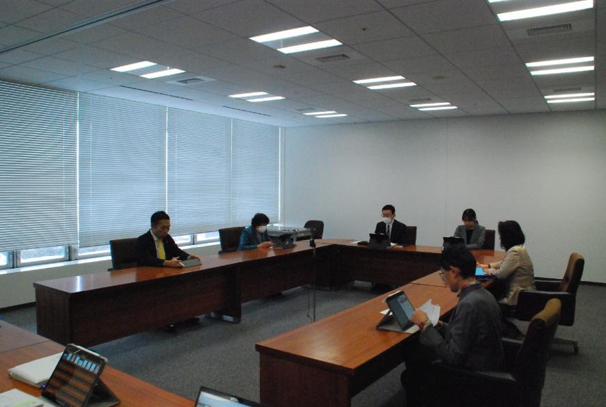 会議室で、コの字型の会議テーブルの席に関係者ら6名が座り、タブレットや書類を見ながら協議を行っている写真