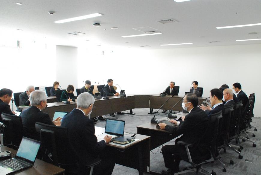 会議室で、ロの字型の会議テーブルの席に女性と男性の関係者らが座り、タブレットや書類を見ながら会議を行っている写真