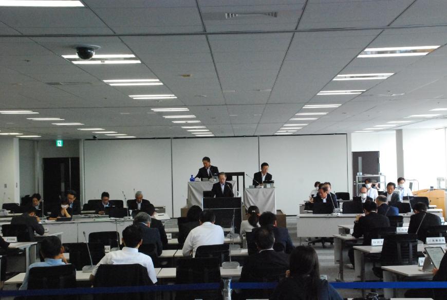 設置された座席に議員さんが座り演台の前に立ち話をする男性の方を見ている写真