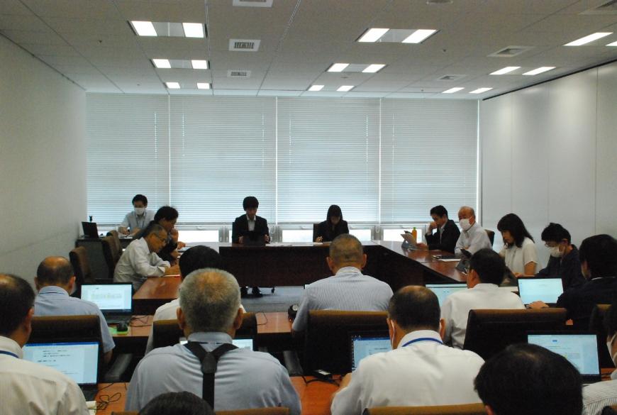 会議室内に設置された長机の席に座ったワイシャツやスーツ姿の参加者が会議をする様子を撮影している写真