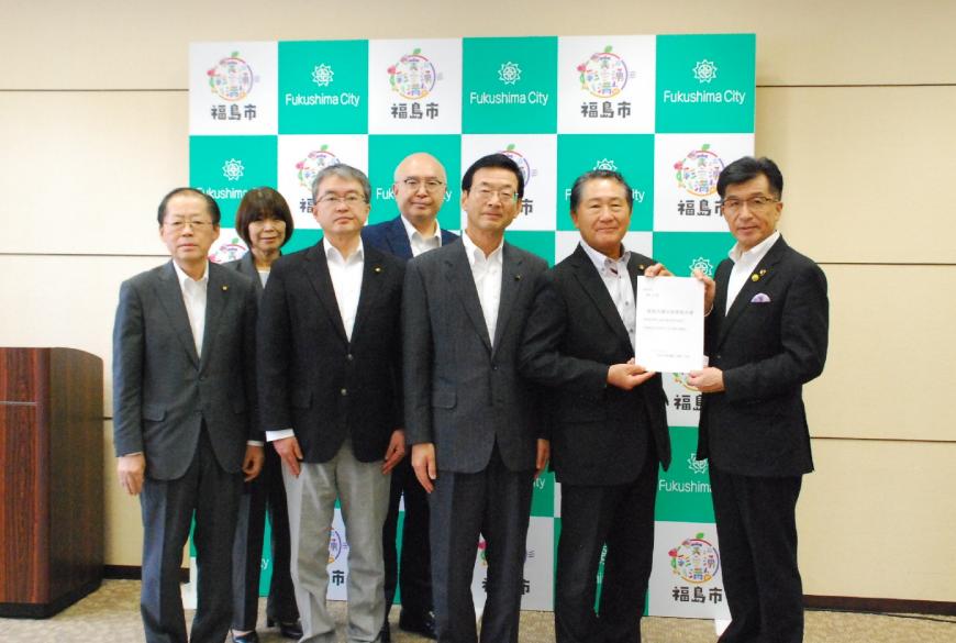 バックパネルの前に常任委員会の方々が立ち、代表の男性と市長が提言書を持ち並んでいる写真
