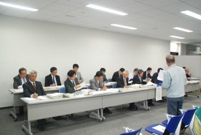 2列に並べられた机と椅子に参加者が座り、手元の資料を見ながら、前方に立つ男性の話を聞いている写真