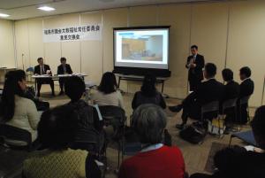 うす暗い室内のスクリーンに資料が投影される横に男性が横に立ち話をしている様子を多くの参加者の方々が聞いている写真