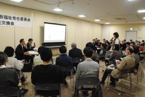 参加者席に座る一人の女性が立ち、マイクを使って前方に座る議員に向けて話をしている写真