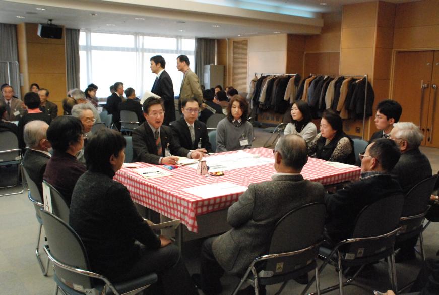 議員の方と参加者たちがテーブルを囲むように座り、その中のスーツ姿の男性が話す様子を聞いている様子の写真