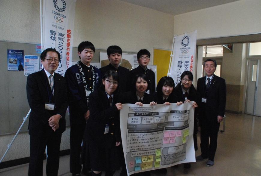 文字や付箋が貼られて模造紙を4人の女子学生が手に持ち、その後ろに男子学生と両サイドにスーツ姿の議員が立ち記念撮影している写真