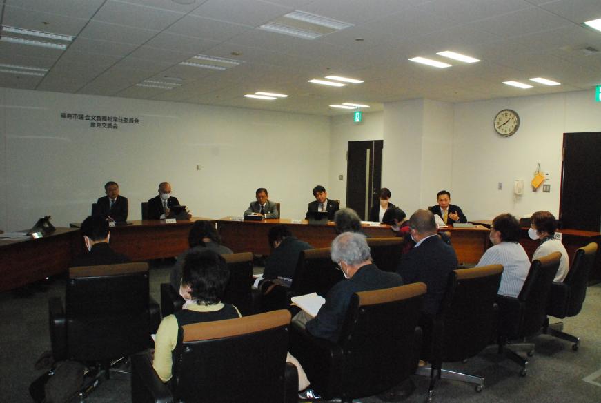 議員席に座る男性の一人が手振りを加え参加者に向けて話をし、それを参加者が聞いている写真