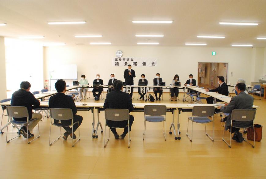 室内にロの字型に設置されたテーブルと椅子に座り資料を見ながら、前方に立つスーツ姿の男性の話を聞いている様子の写真