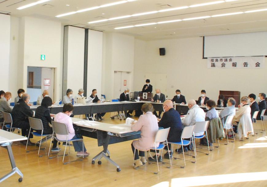 室内にロの字型に設置されたテーブルと椅子に座り資料を見ながら司会者の話を聞いている様子の写真