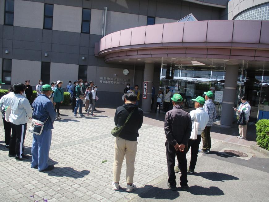建物の入口前に、参加者と緑色の帽子をかぶった関係者が集まり輪になって立っている写真