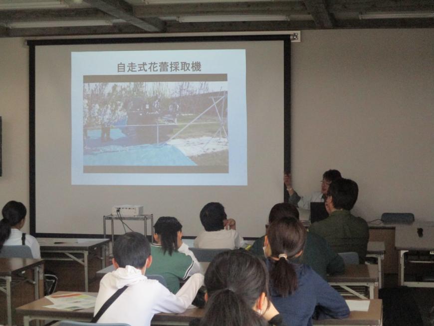 前方の大きなスクリーンに映し出された資料を長机の席に座って見ている参加者たちの写真