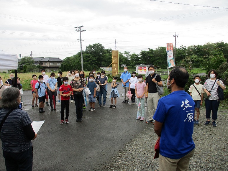 屋外に集まった親子連れなどの参加者の前で、青いTシャツを着た男性が挨拶をしている写真