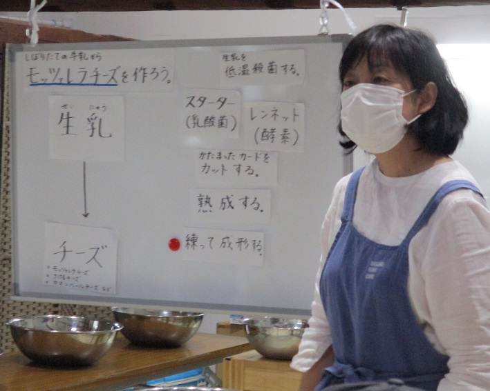 モッツァレラチーズの作り方が記されたホワイトボードの前に青いエプロンをした女性が立っている写真