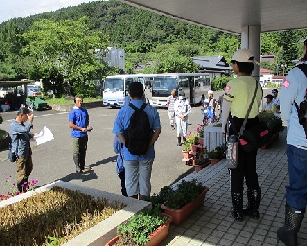 建物の入り口前に参加者や関係者が集まっており、駐車場には2台のマイクロバスが停まっている写真